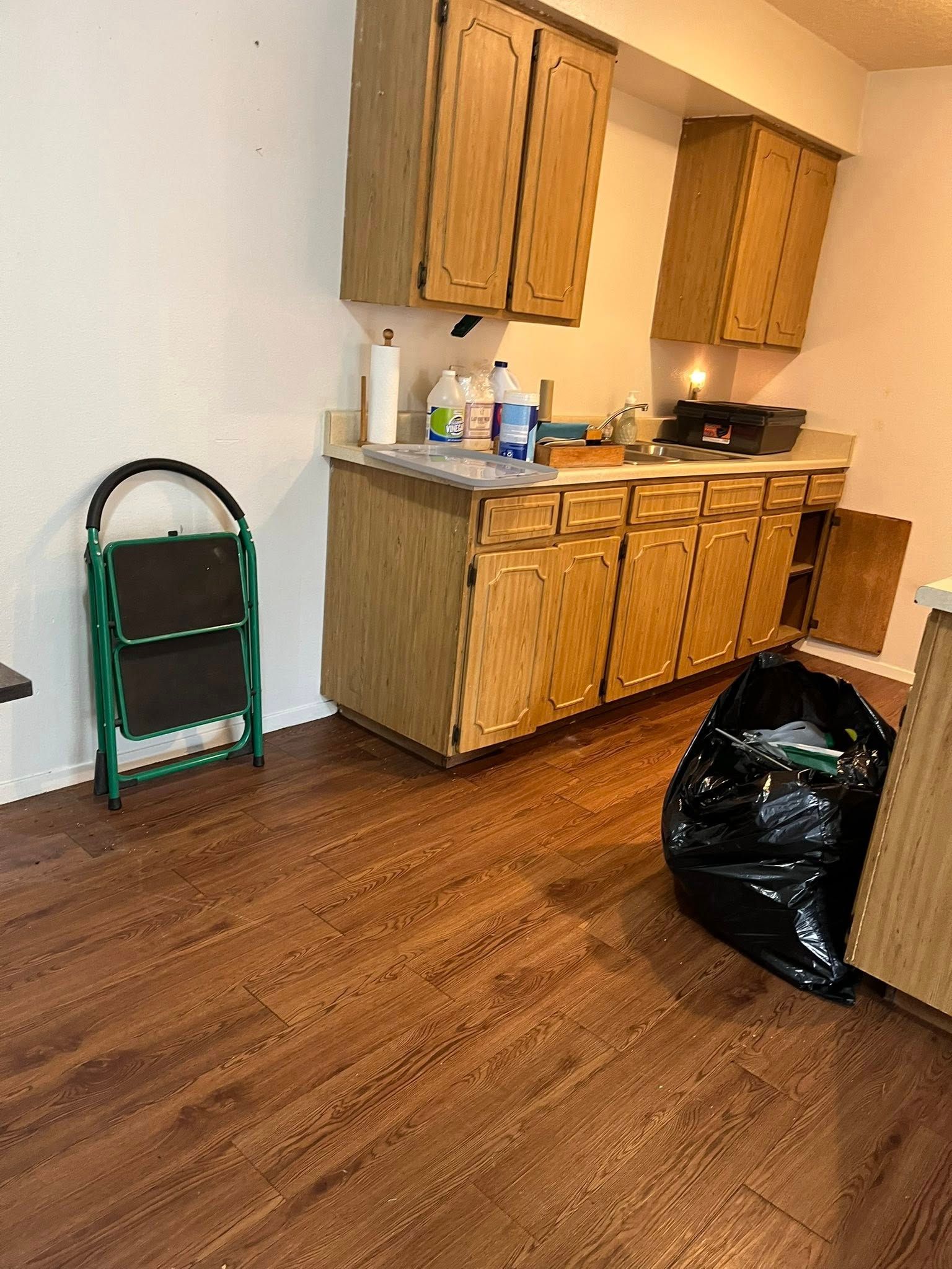 Small kitchen with wood cabinets, step stool, trash bag, and wood-look flooring.