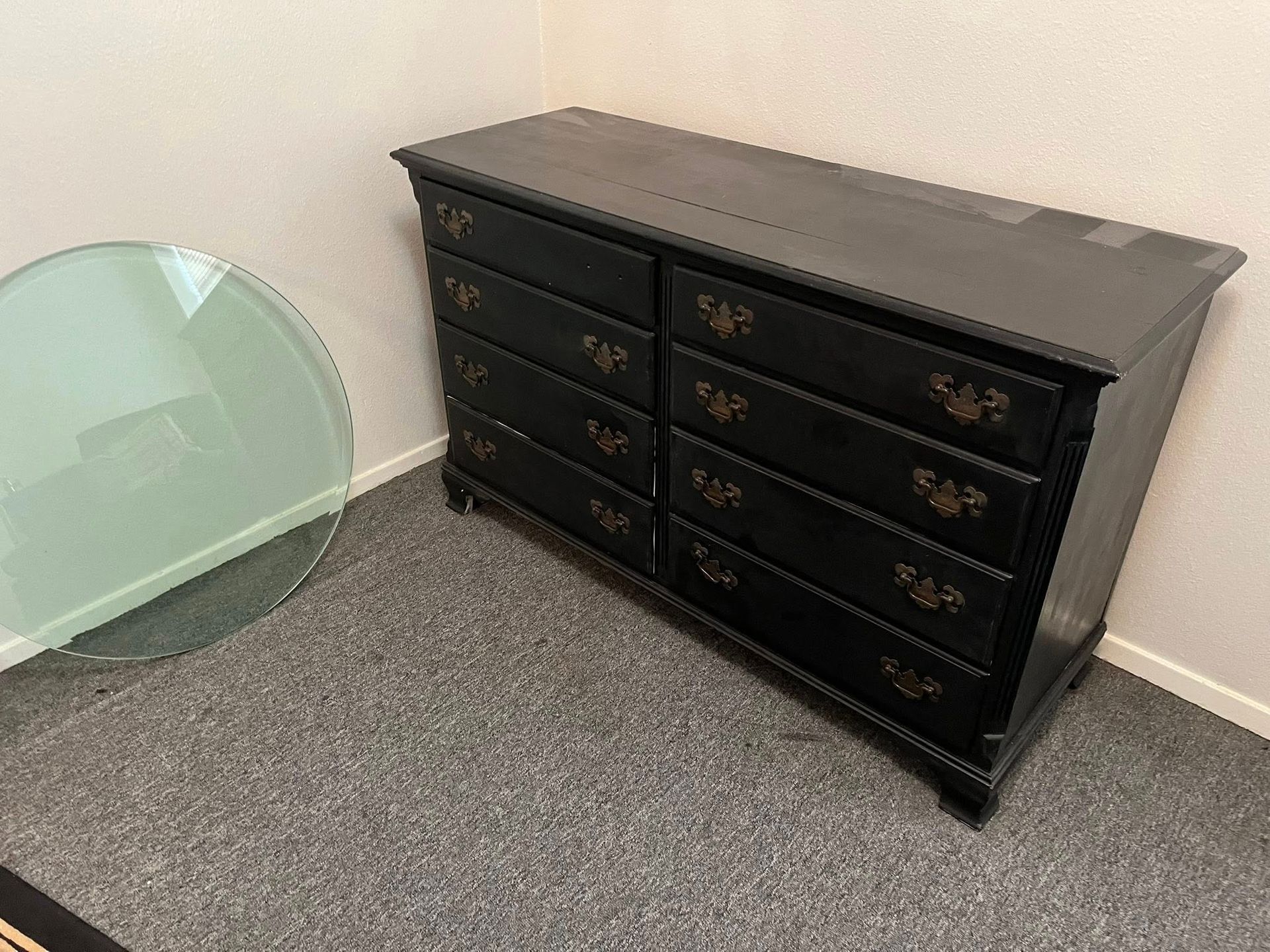 Black dresser with eight drawers in room with gray carpet and round glass table.