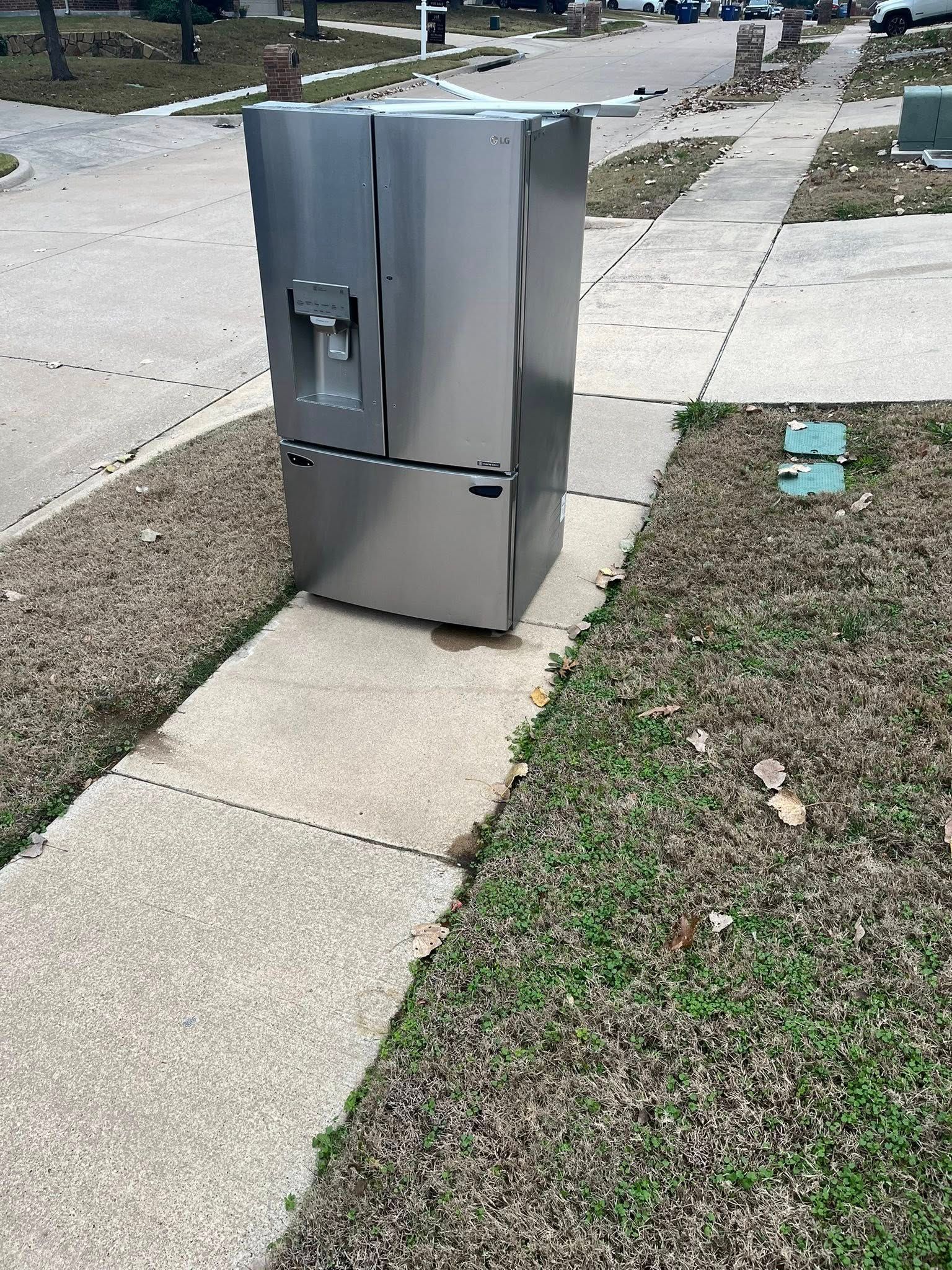 Stainless steel refrigerator on a sidewalk next to a grassy area in a neighborhood.