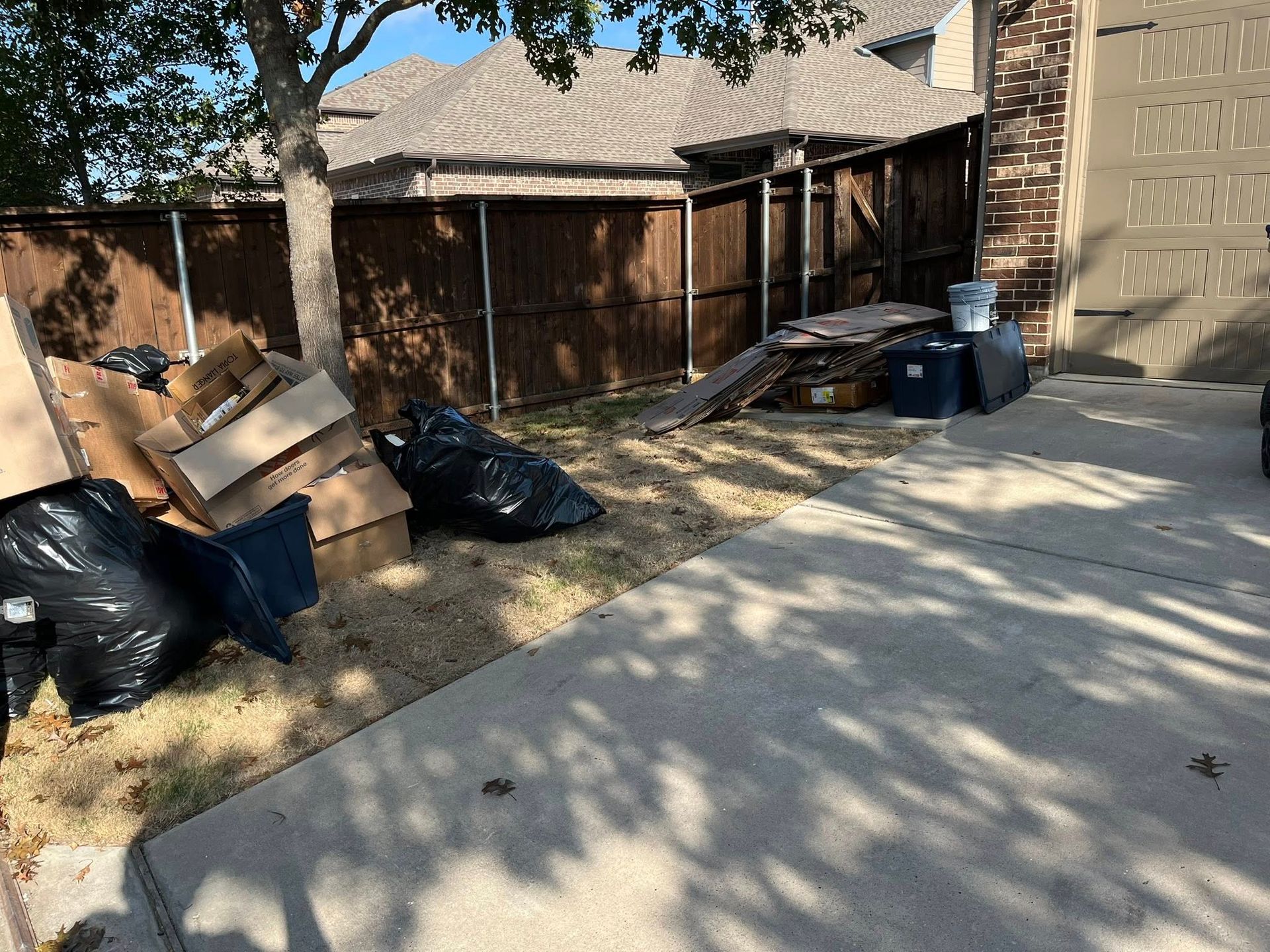 Yard with clutter: cardboard boxes, black trash bags, blue bins, and lumber by a wooden fence.