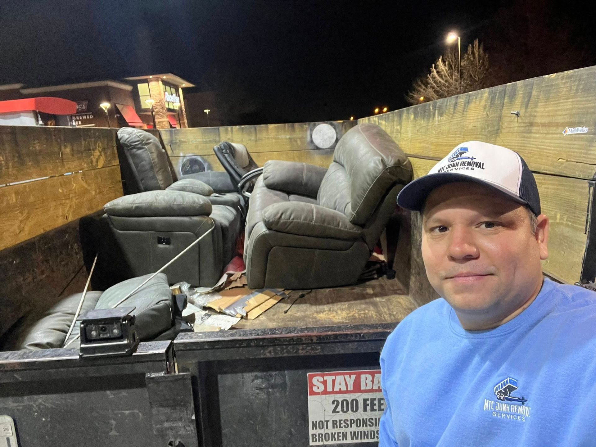 Man in a cap posing beside a truck bed filled with old chairs and furniture at night