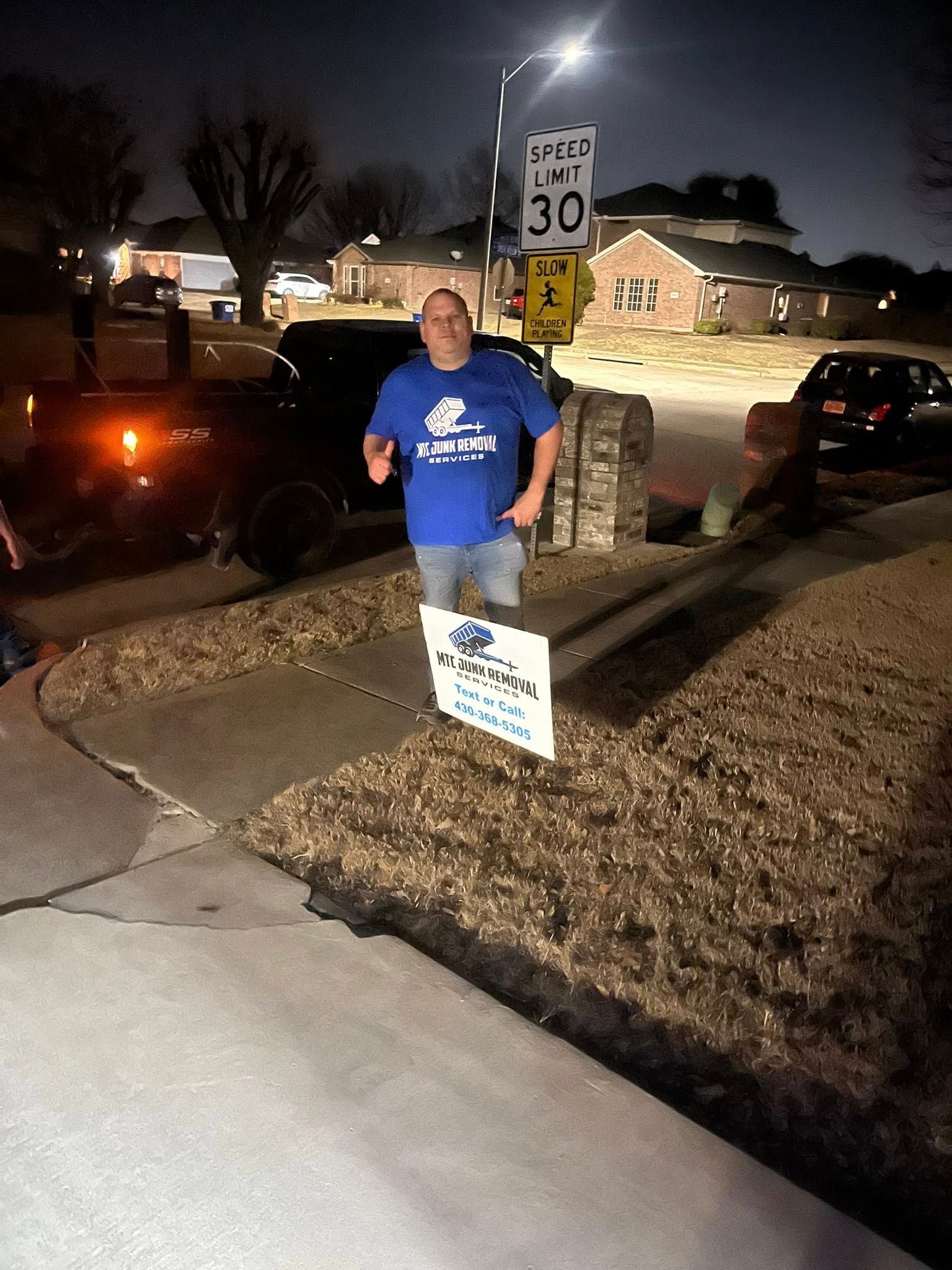 Person in a blue shirt standing on a sidewalk at night, holding signs near parked cars and a speed limit sign