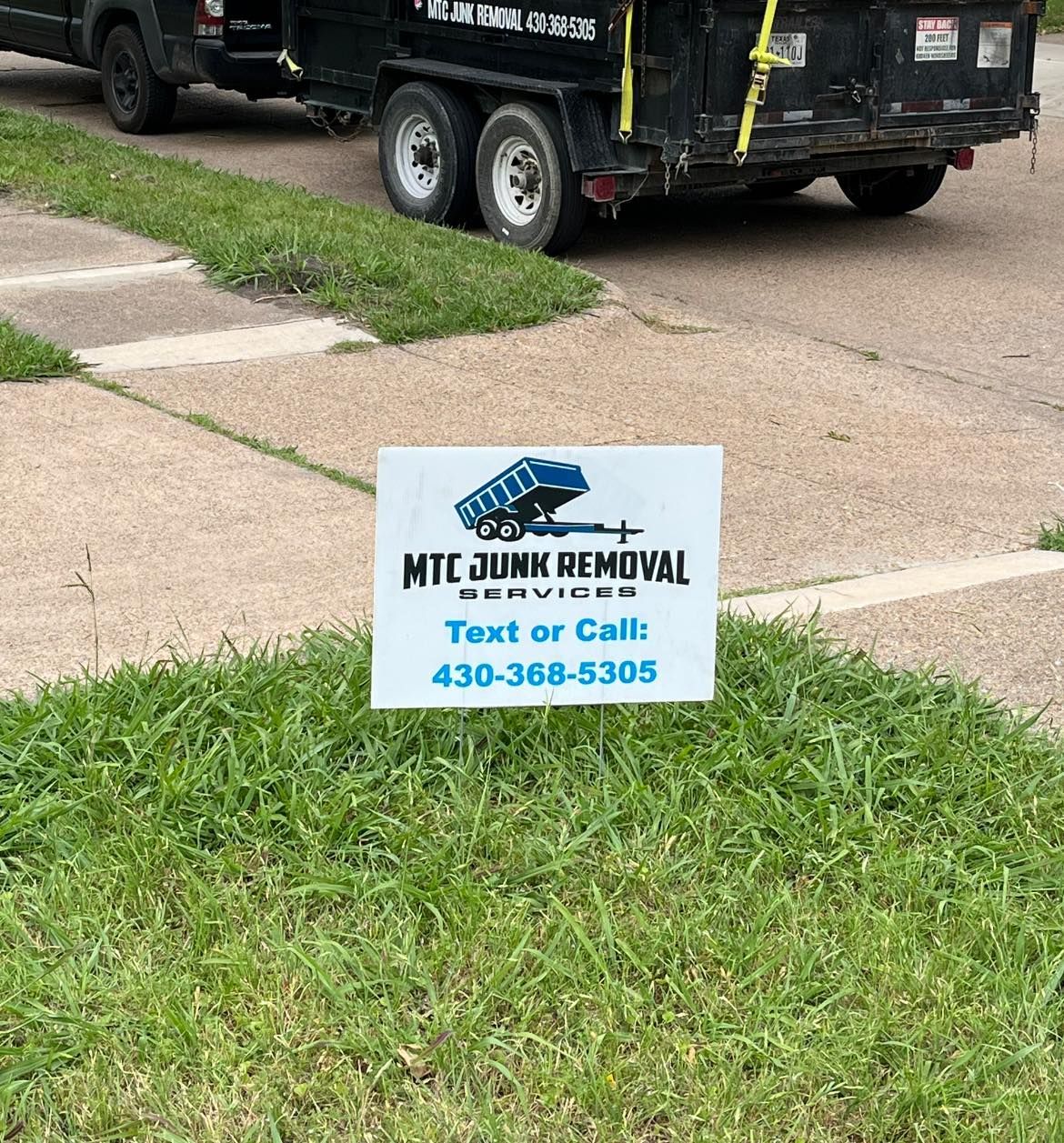 HMT Junk Removal yard sign on grass beside driveway, with black dump trailer in background