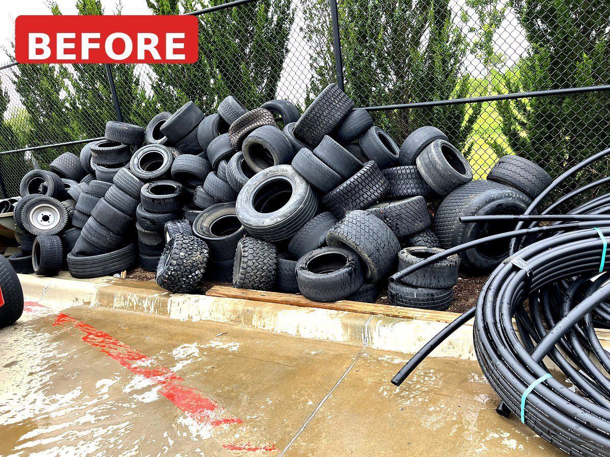 Pile of discarded tires and black hoses behind a chain-link fence, with “BEFORE” label and wet pavement in front