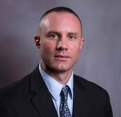 Man in a suit and tie, looking forward, with a slight smile, against a grey background.