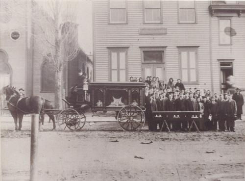 Horse-drawn hearse, men in suits, and mourners pose outside a building, suggesting a funeral.
