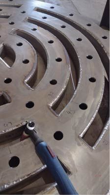 Soldadura sobre placa metálica de gran tamaño con cortes en forma de arco y agujeros perforados; antorcha de soldadura azul visible.