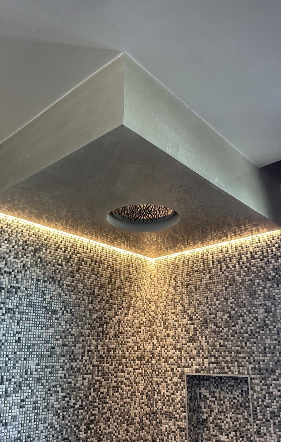 Un angolo doccia con soffitto texturizzato, soffione a pioggia circolare e illuminazione a LED a incasso dai toni caldi, sullo sfondo di piastrelle a mosaico.