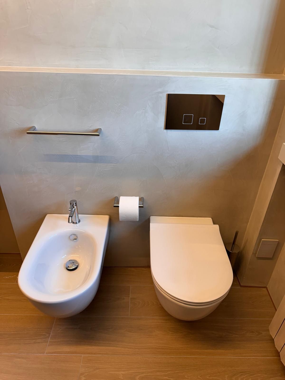Un bidet a parete e un WC con pulsante di scarico sulla parete sovrastante, su uno sfondo grigio chiaro.