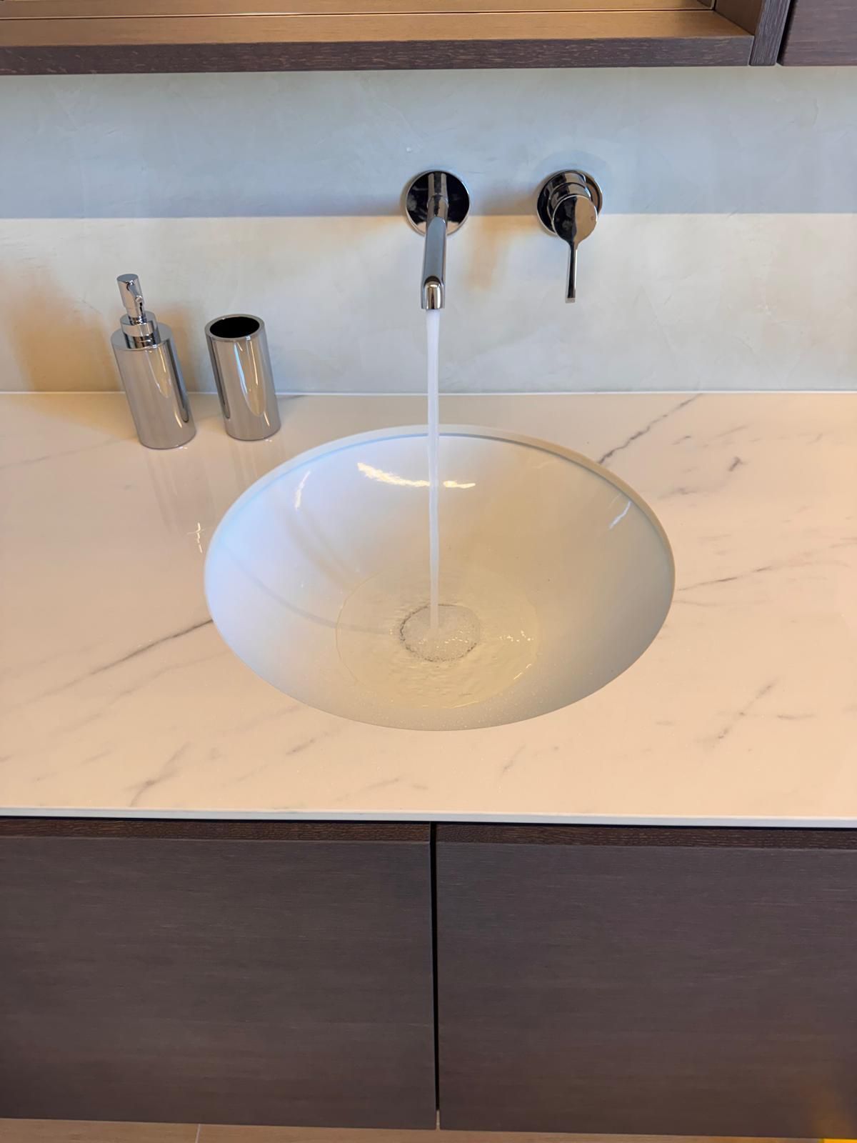 Un mobile da bagno con piano in marmo bianco, lavabo rotondo e rubinetto a parete cromato con acqua corrente.