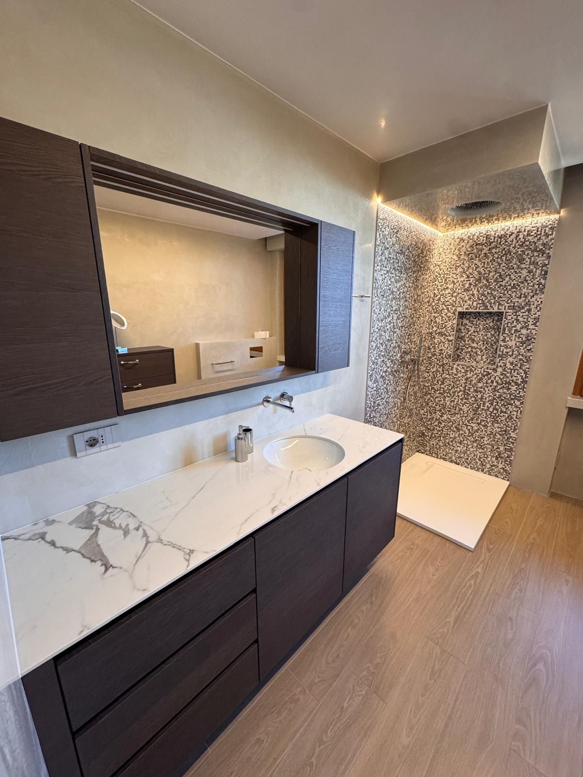 Bagno moderno con mobile lavabo in marmo bianco, mobili in legno scuro, un grande specchio e una cabina doccia con piastrelle a mosaico.