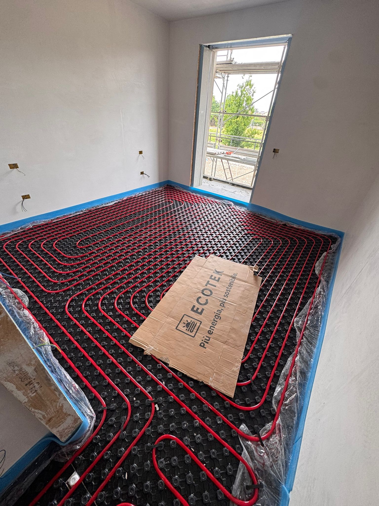 All'interno di una stanza non rifinita con un'ampia finestra, sono stati installati tubi rossi per il riscaldamento a pavimento, disposti a griglia su una base scura.