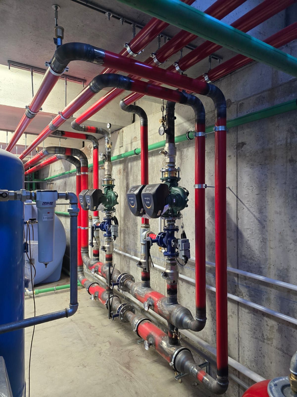 Tubature industriali rosse e grigie e pompe di circolazione installate lungo una parete di cemento in un locale tecnico.