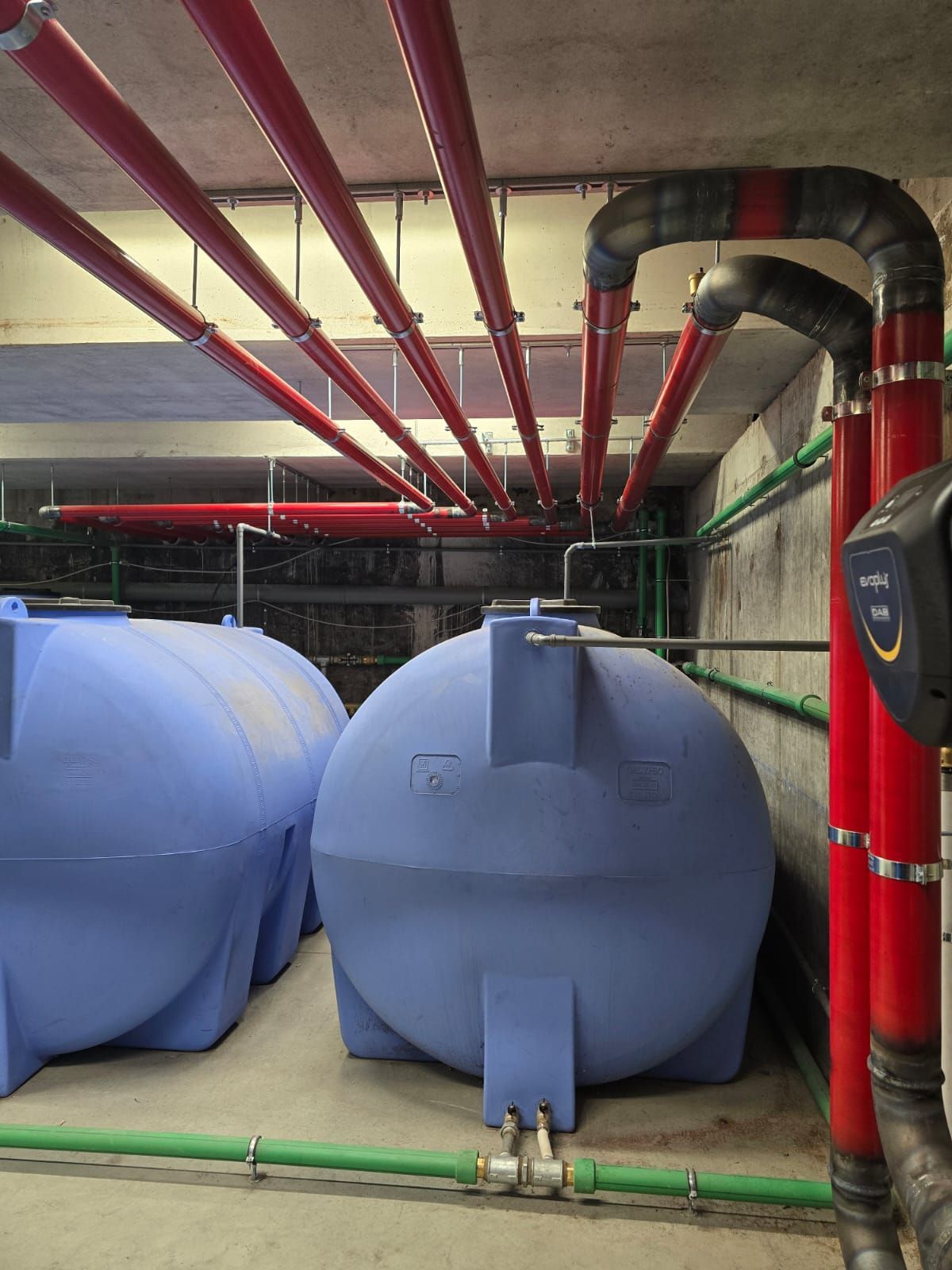 Due grandi serbatoi d'acqua blu in un locale seminterrato, situati sotto una rete di tubi rossi aerei e tubi verdi a parete.