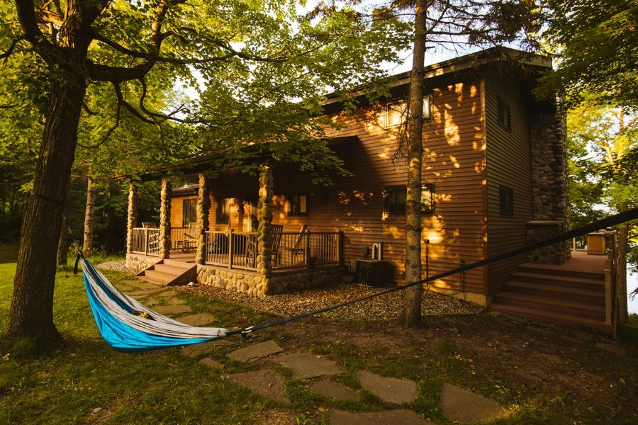 A hammock is sitting in front of a wooden house surrounded by trees.