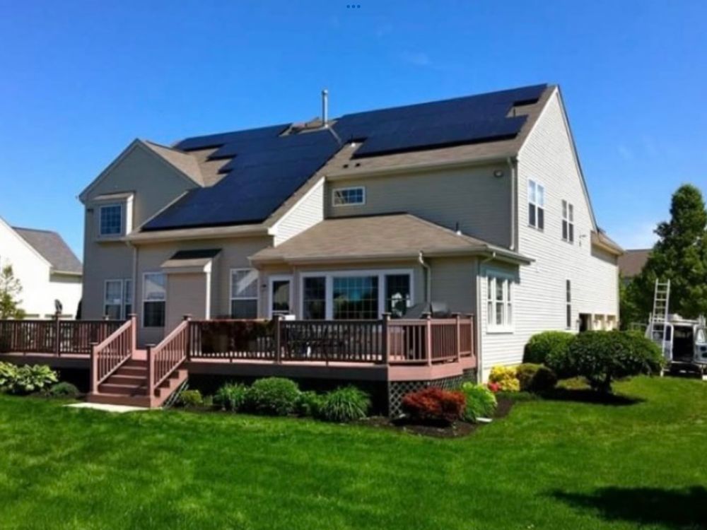 Solar panels on a beige house with a wooden deck and a green lawn. Blue sky overhead.