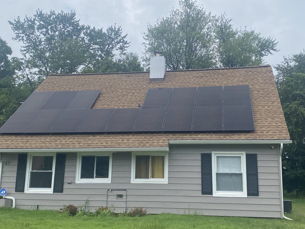 Gray house with solar panels on the roof, surrounded by trees, with a cloudy sky.