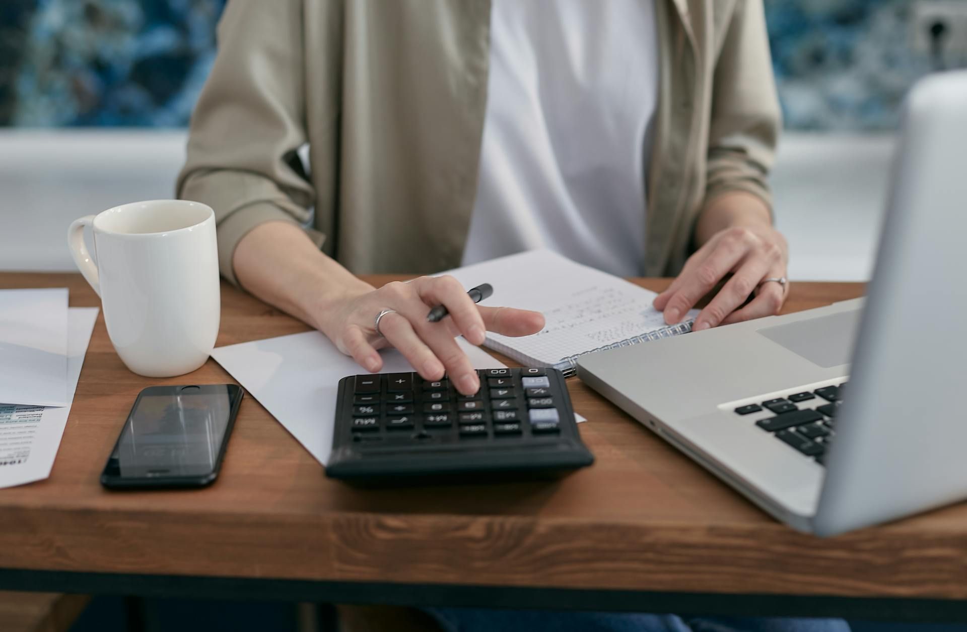 A woman is sitting at a desk using a calculator and a laptop.