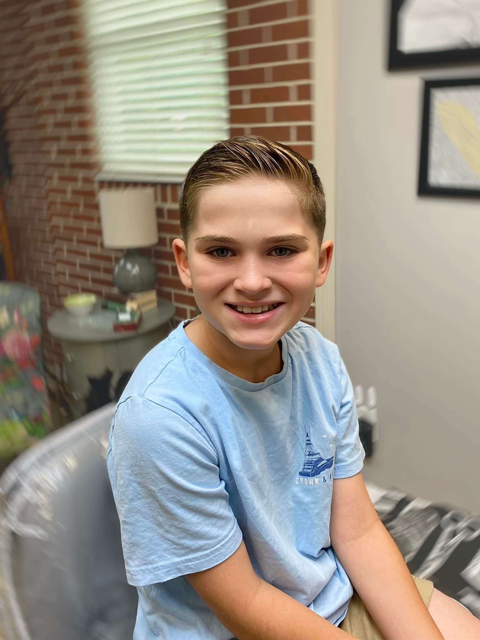 A young boy in a blue shirt is sitting in a chair and smiling.