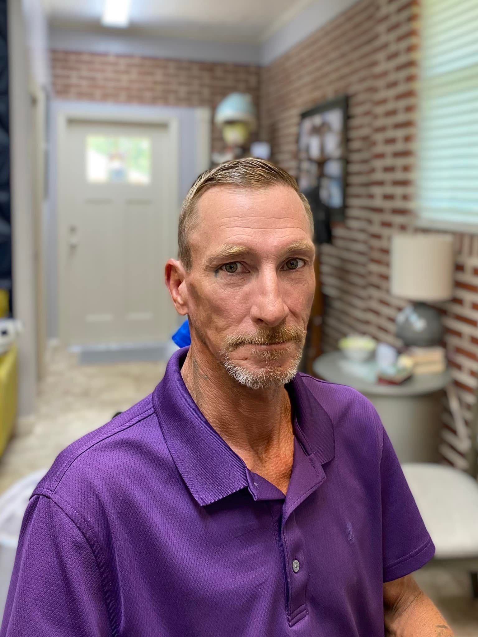 A man in a purple shirt is standing in a room with a brick wall.