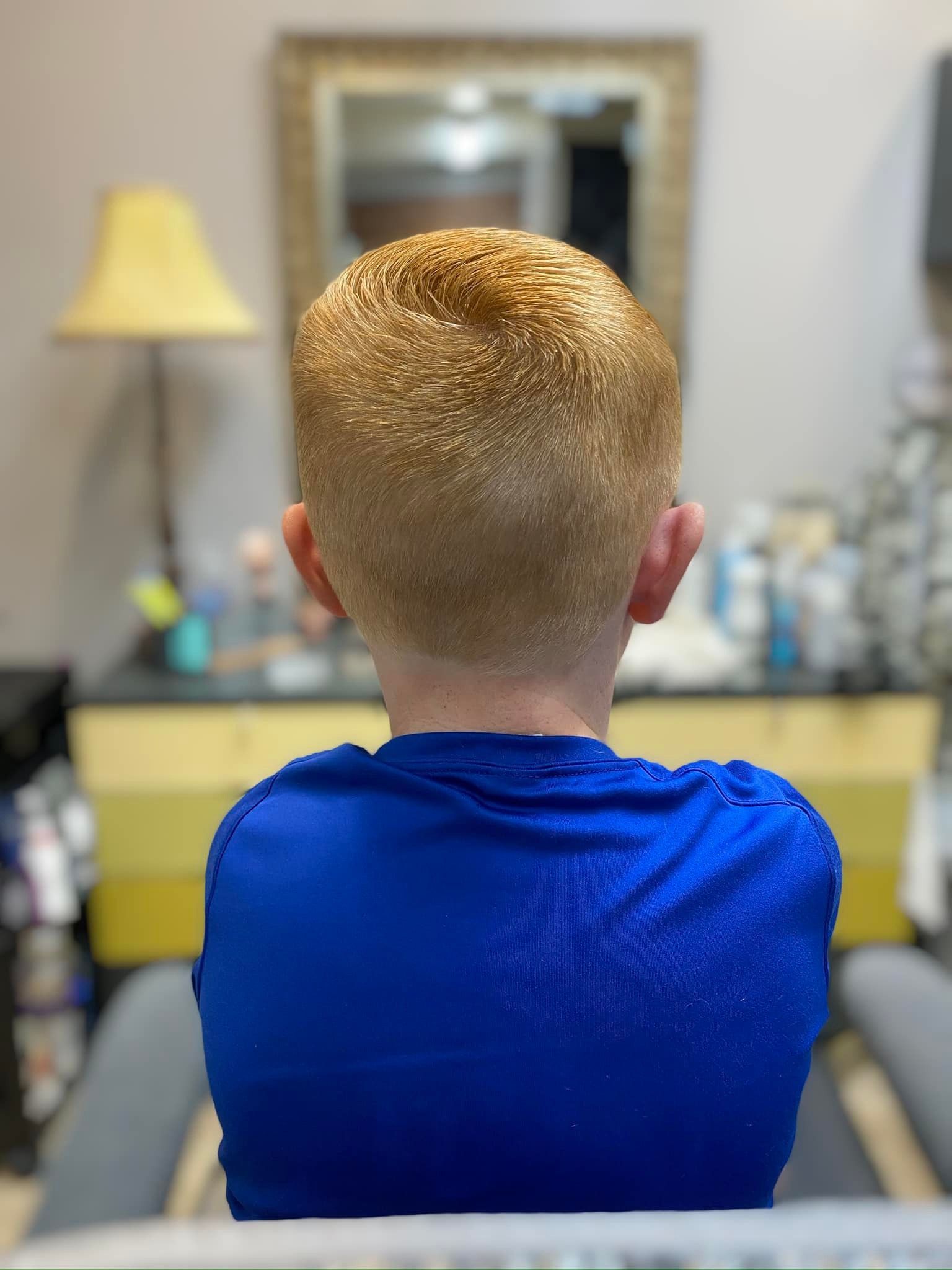 A young boy in a blue shirt is sitting in a chair in front of a mirror.