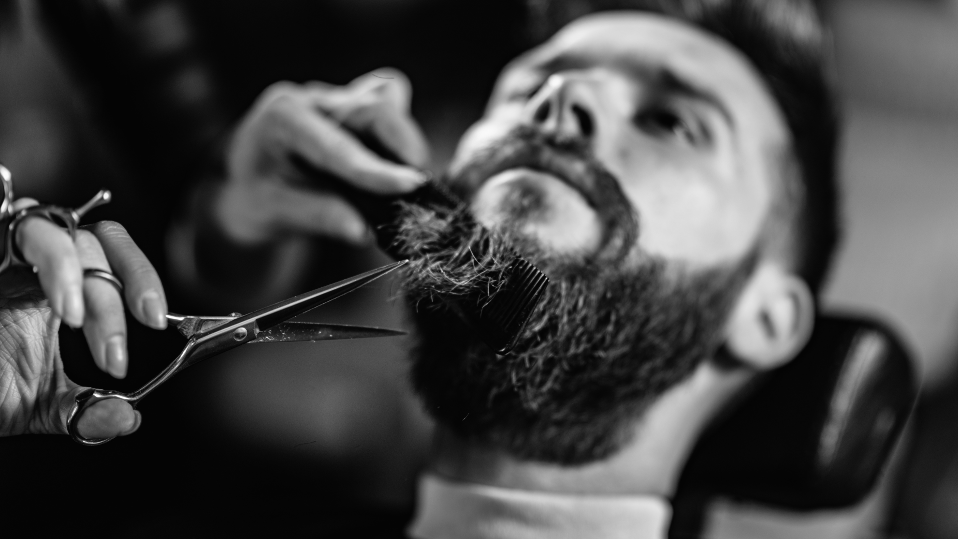 A man is getting his beard cut by a barber with scissors.