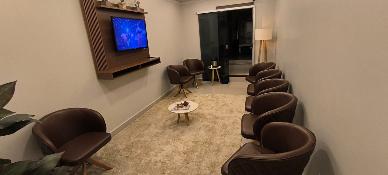 Uma sala de espera com cadeiras marrons, uma mesinha e uma TV na parede.