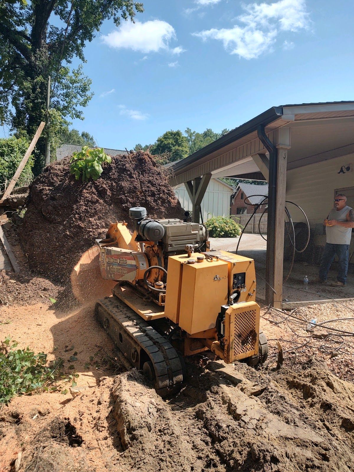 A yellow bulldozer is cutting a tree stump in front of a garage.