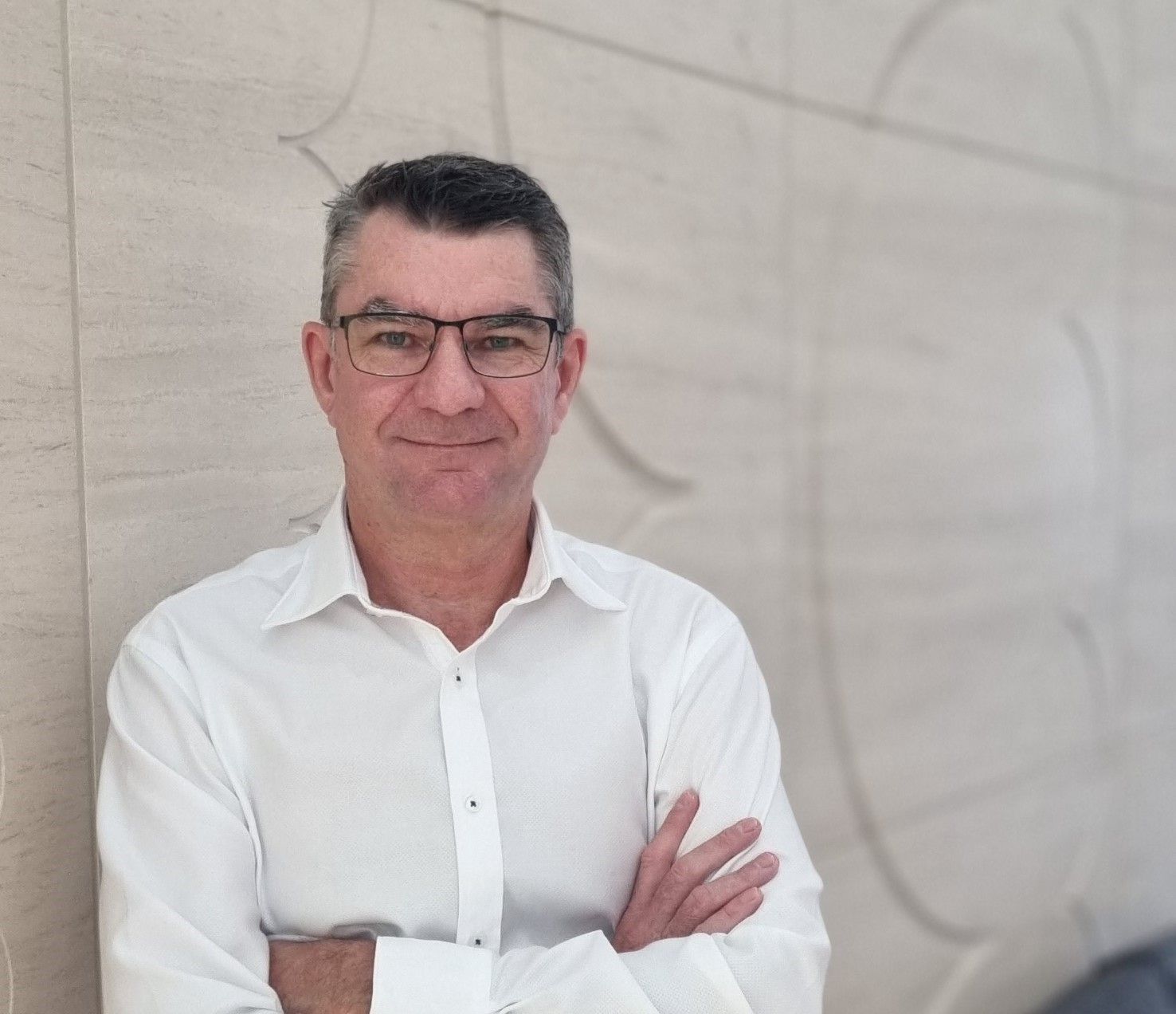 A man wearing glasses and a white shirt is leaning against a wall with his arms crossed.