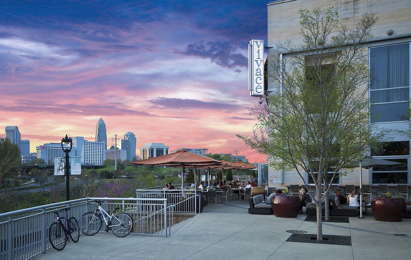 Restaurant patio with cityscape at sunset. People dine at tables under umbrellas. Bicycles lean on a railing.