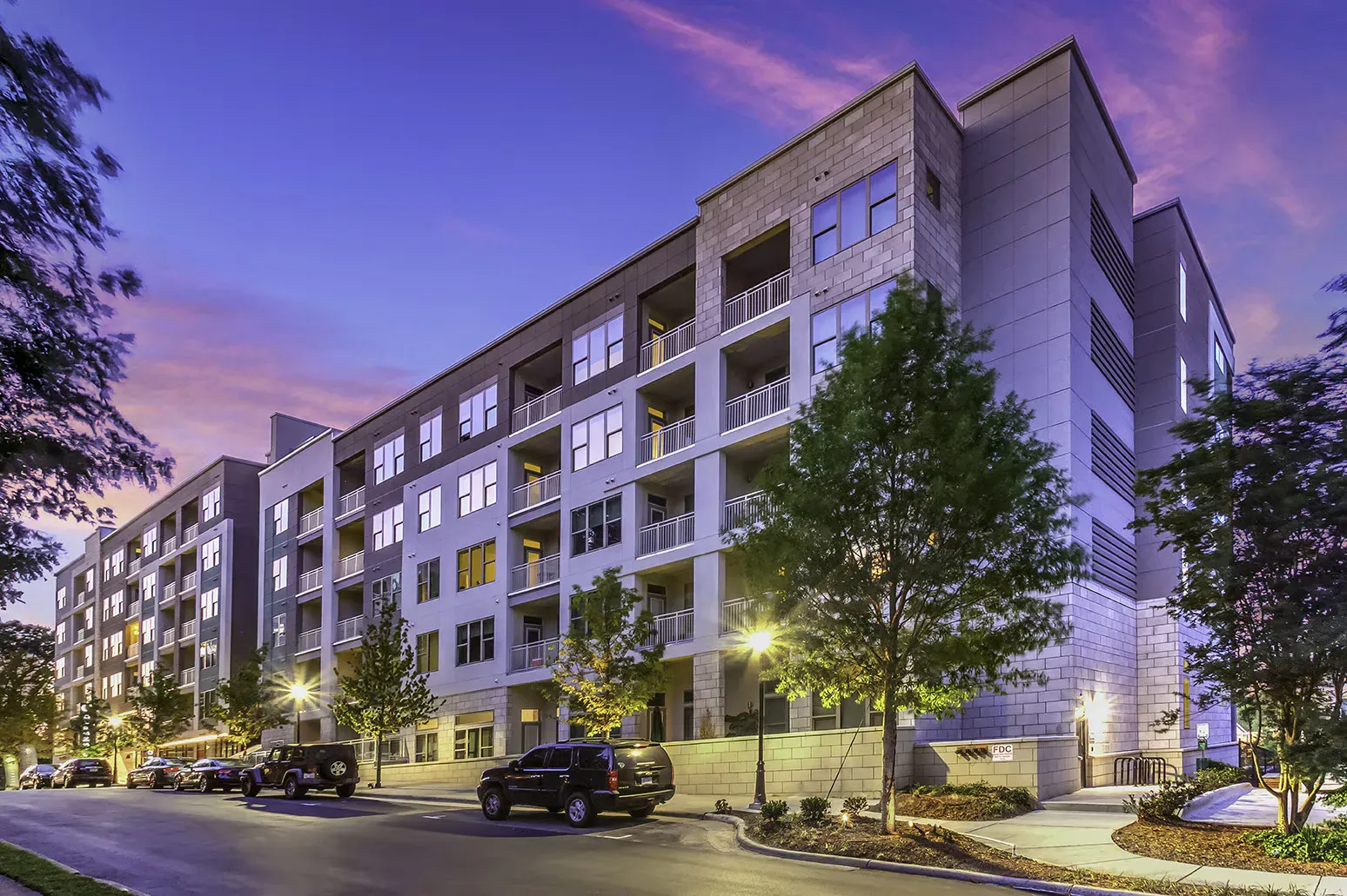 Multi-story apartment building at dusk with street parking and landscaping.