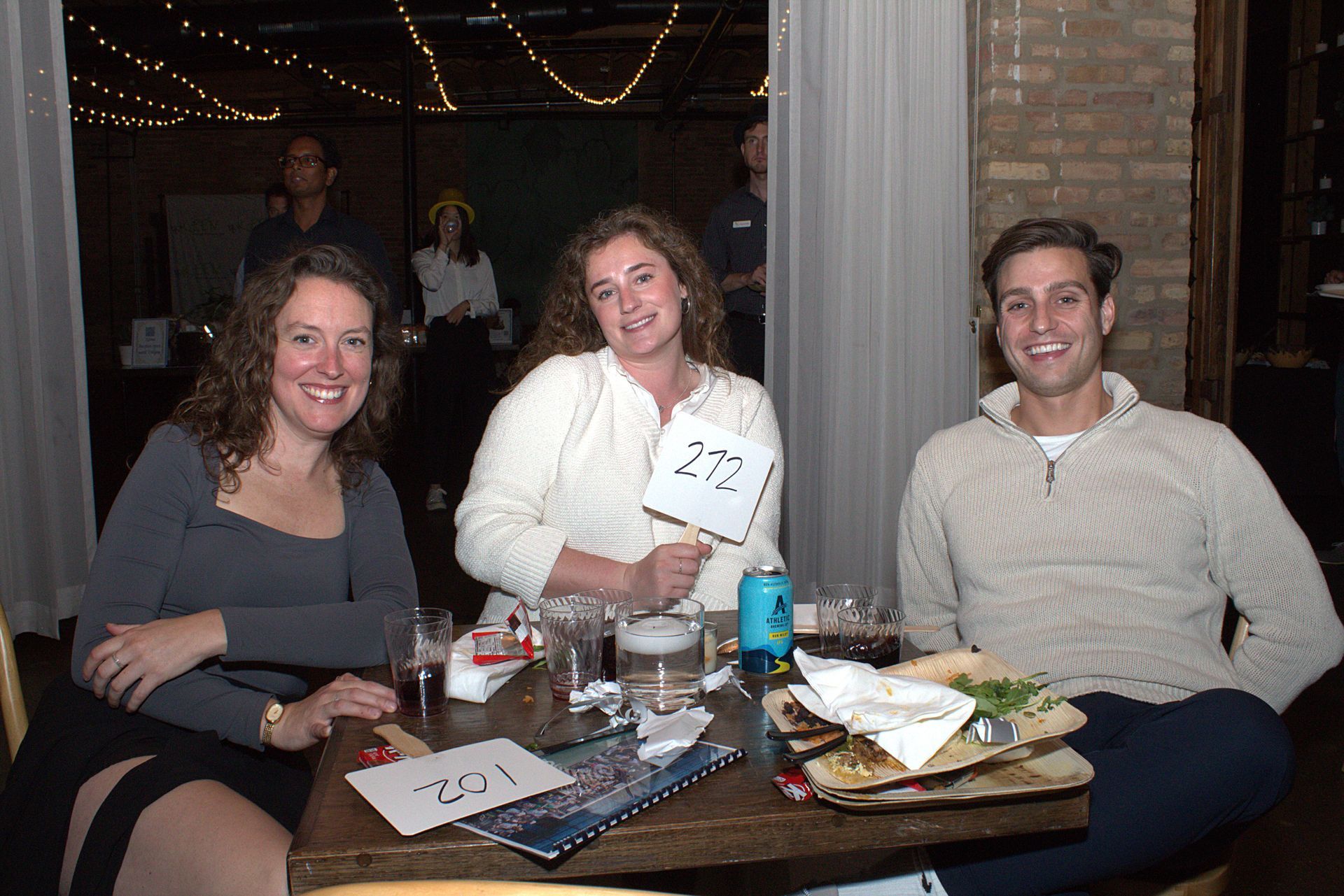 Event attendees eat and drink at the table, smiling for the camera and holding up a bid number