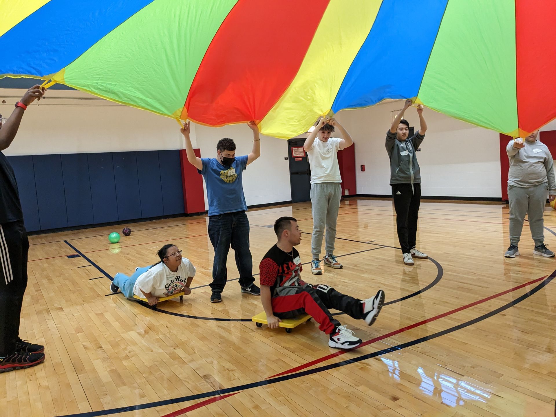 A group of youth plays with a parachute. Two youth with down syndrome go under the parachute on scooters