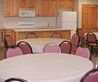Dining room with round tables covered in white linens, pink chairs, and a kitchenette in the background.