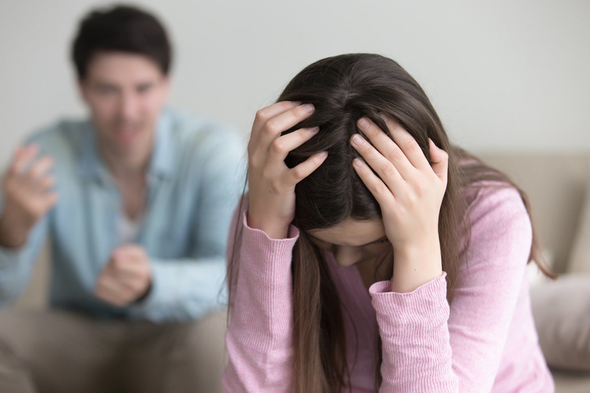 A man is sitting on a couch yelling at a woman who is covering her face with her hands.