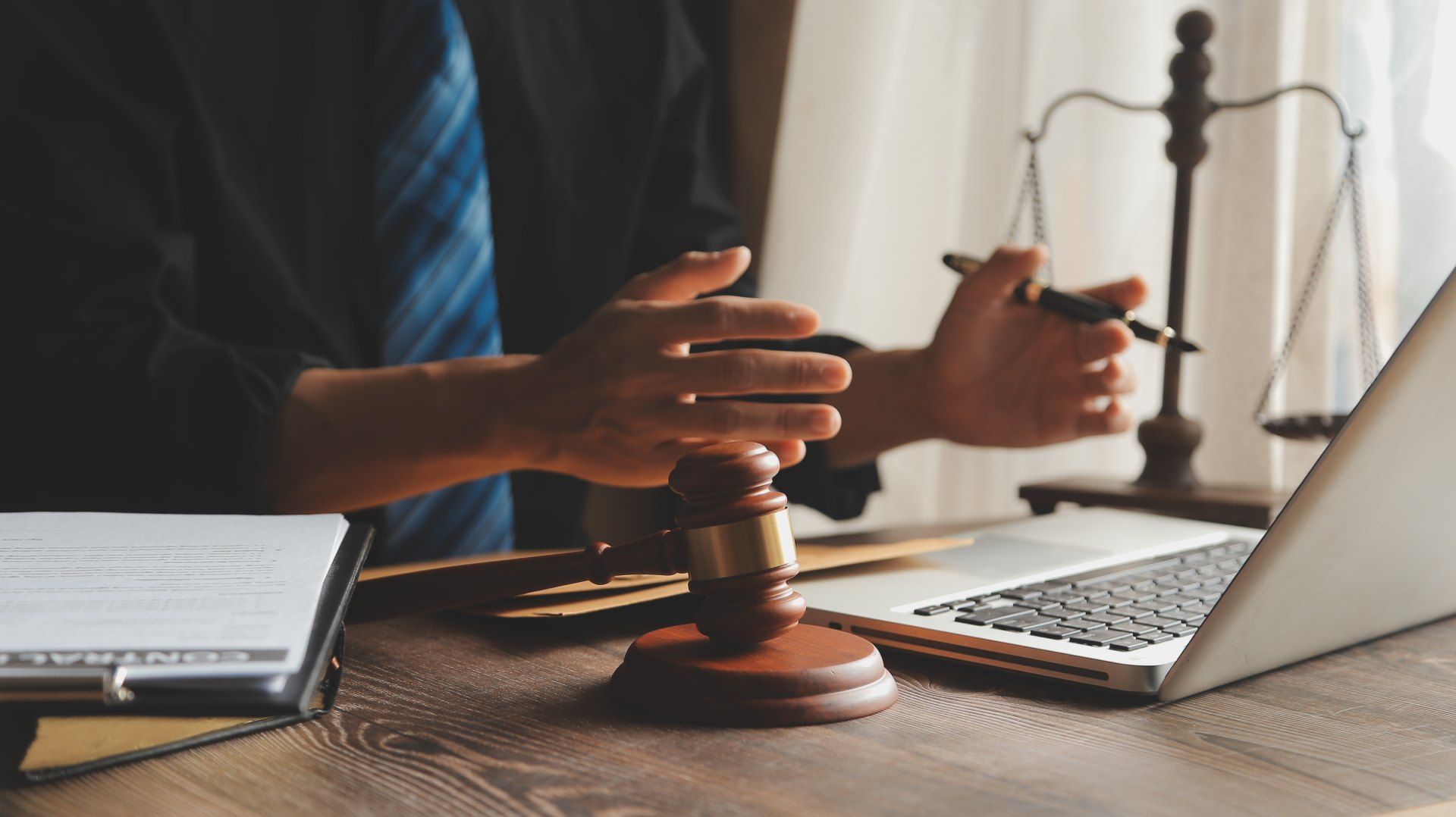 Person in suit with gavel, laptop, and scales on desk, gesturing with pen.