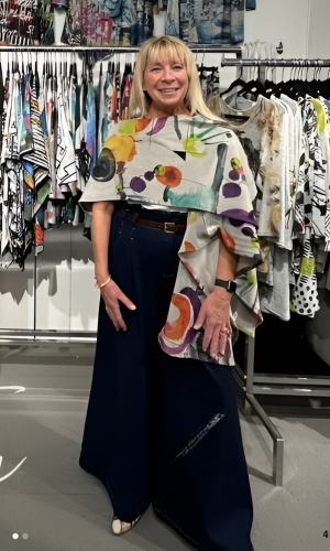 Woman in a boutique wearing a colorful floral wrap over wide-leg blue pants. She's smiling, surrounded by clothing racks.