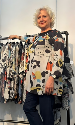 Woman with curly blonde hair, smiling, wearing a patterned tunic and dark pants.