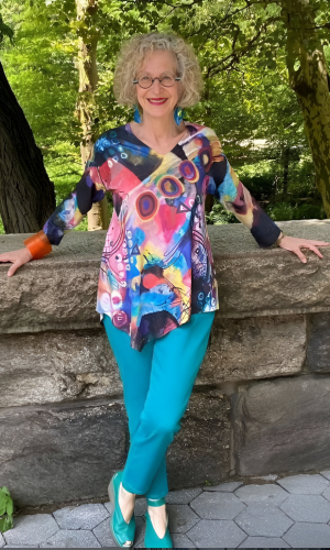 Woman in turquoise pants and colorful top stands against a stone wall in a park, smiling.