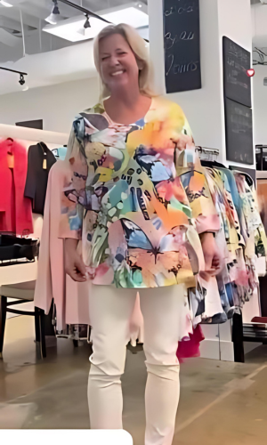 Woman in a colorful butterfly-print top and white pants smiling in a clothing store.