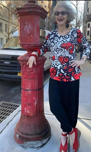 Woman in colorful top and pants smiles next to a red vintage mailbox on a city street.