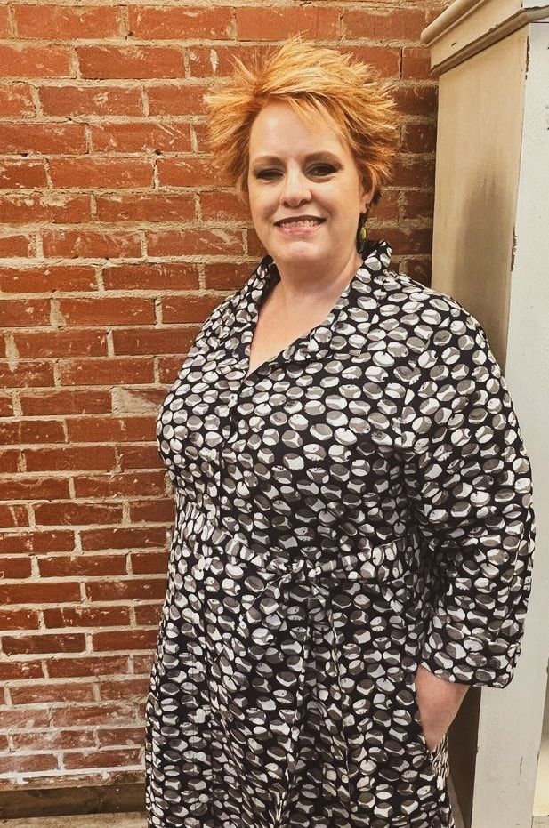 Woman with short, spiky red hair, smiling, wearing a black and white patterned dress. Leaning against a brick wall.