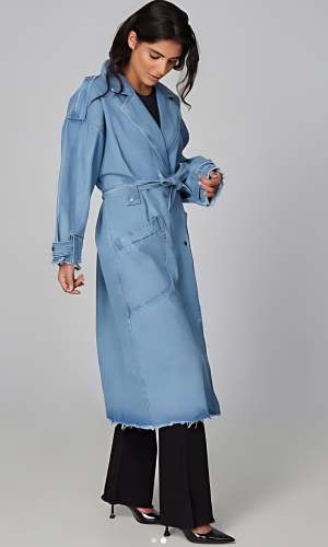 Woman in denim trench coat and black pants, standing in a studio. Coat has a tie belt, frayed edges.