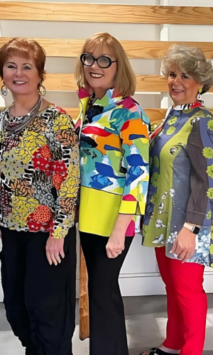 Three women in colorful blouses and black/red pants posing in front of a wooden structure; one smiles broadly.