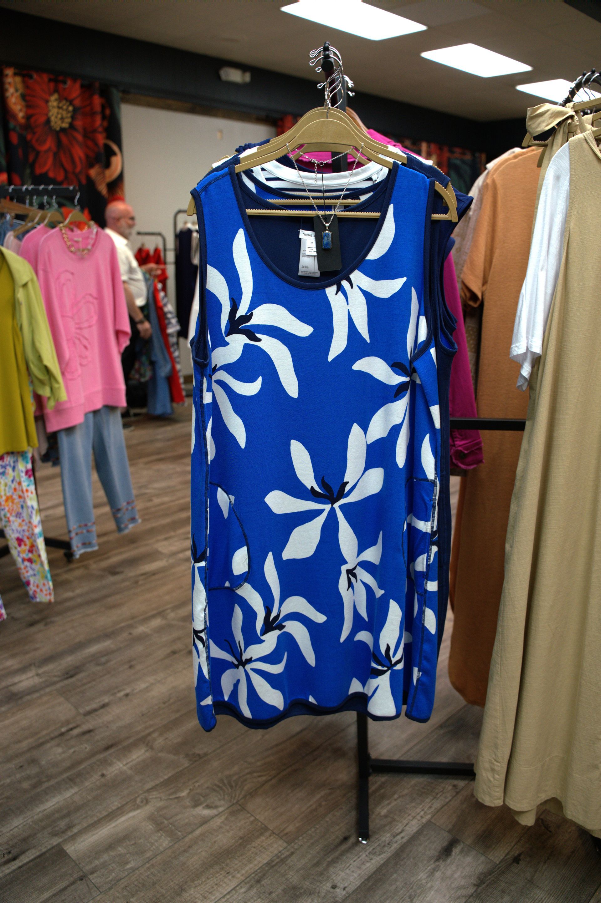 A blue dress with white floral patterns hangs on a rack inside a clothing store. Other garments are visible around it.