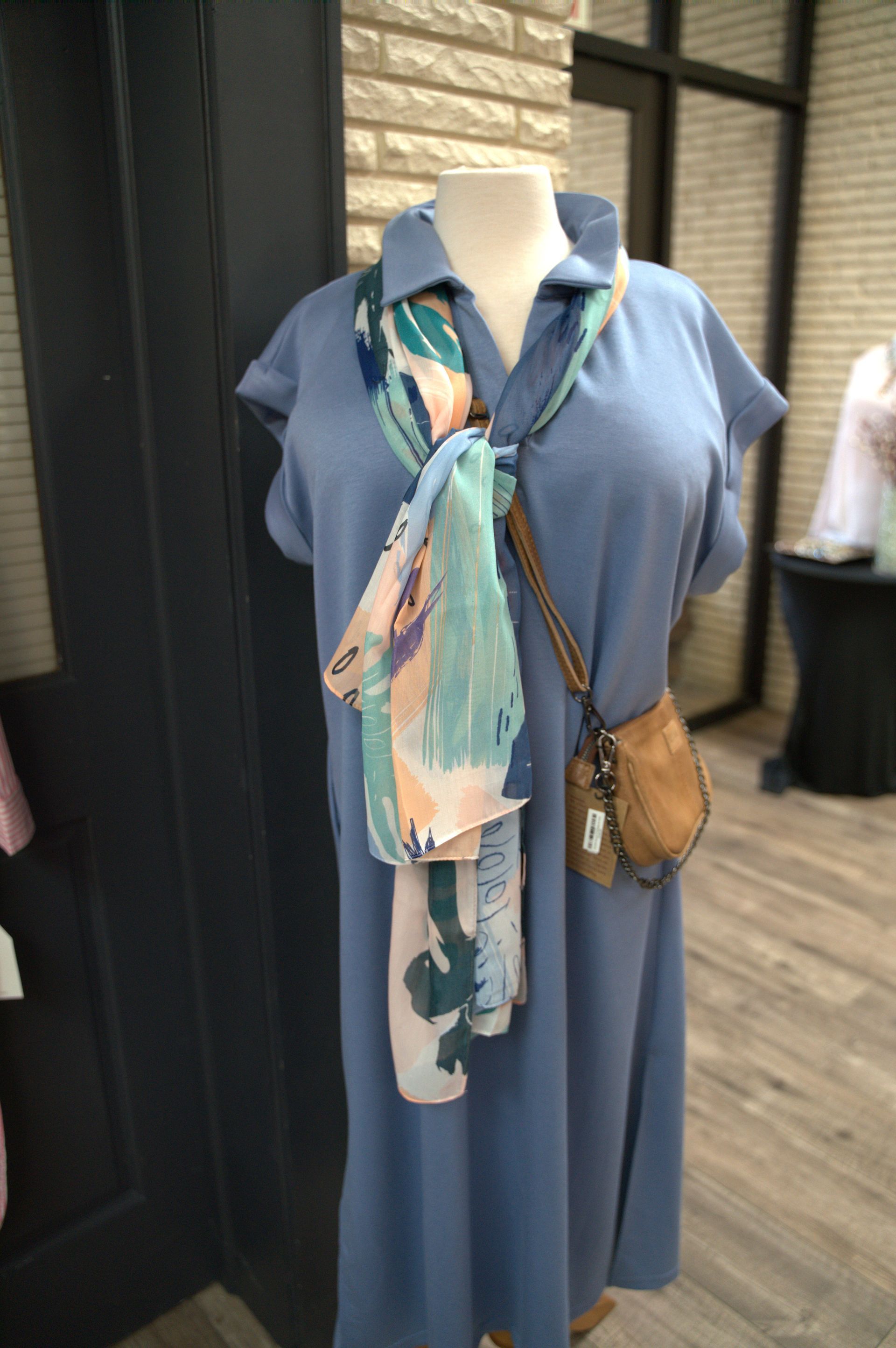 A mannequin dressed in a blue collared dress, patterned scarf, and small crossbody bag.