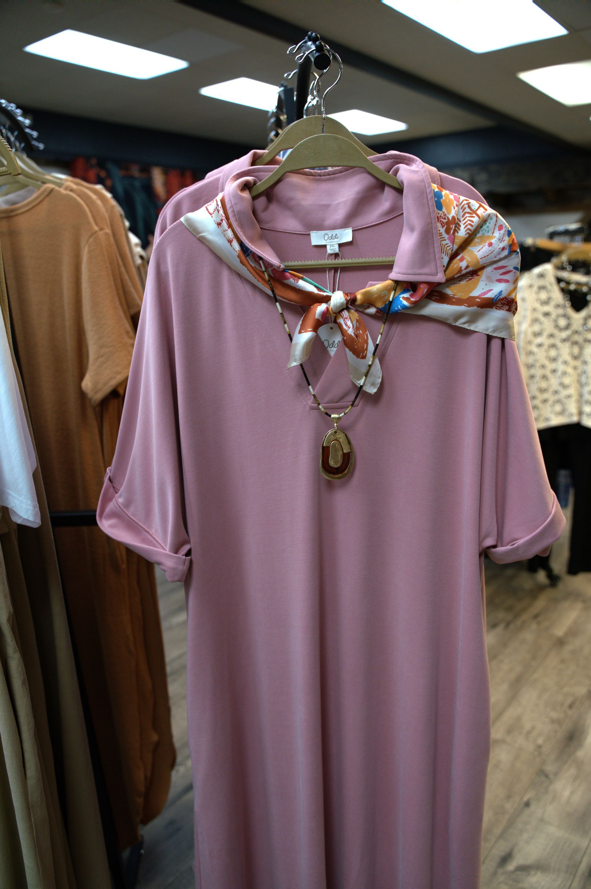 A pink midi dress with rolled sleeves is displayed on a rack, adorned with a patterned scarf and pendant necklace.