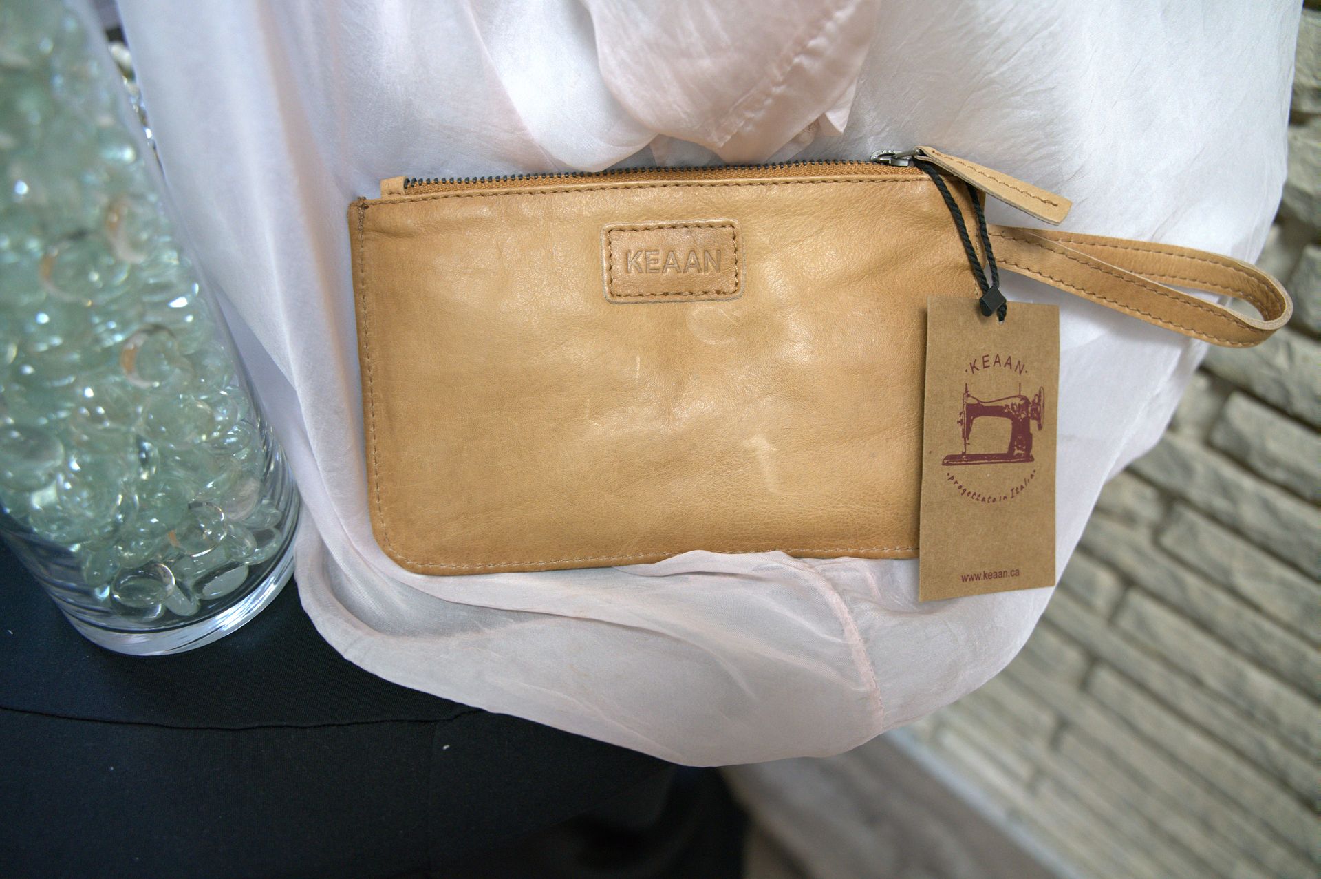 Tan leather clutch with a wrist strap and a tag, set against a sheer fabric and a stone wall.