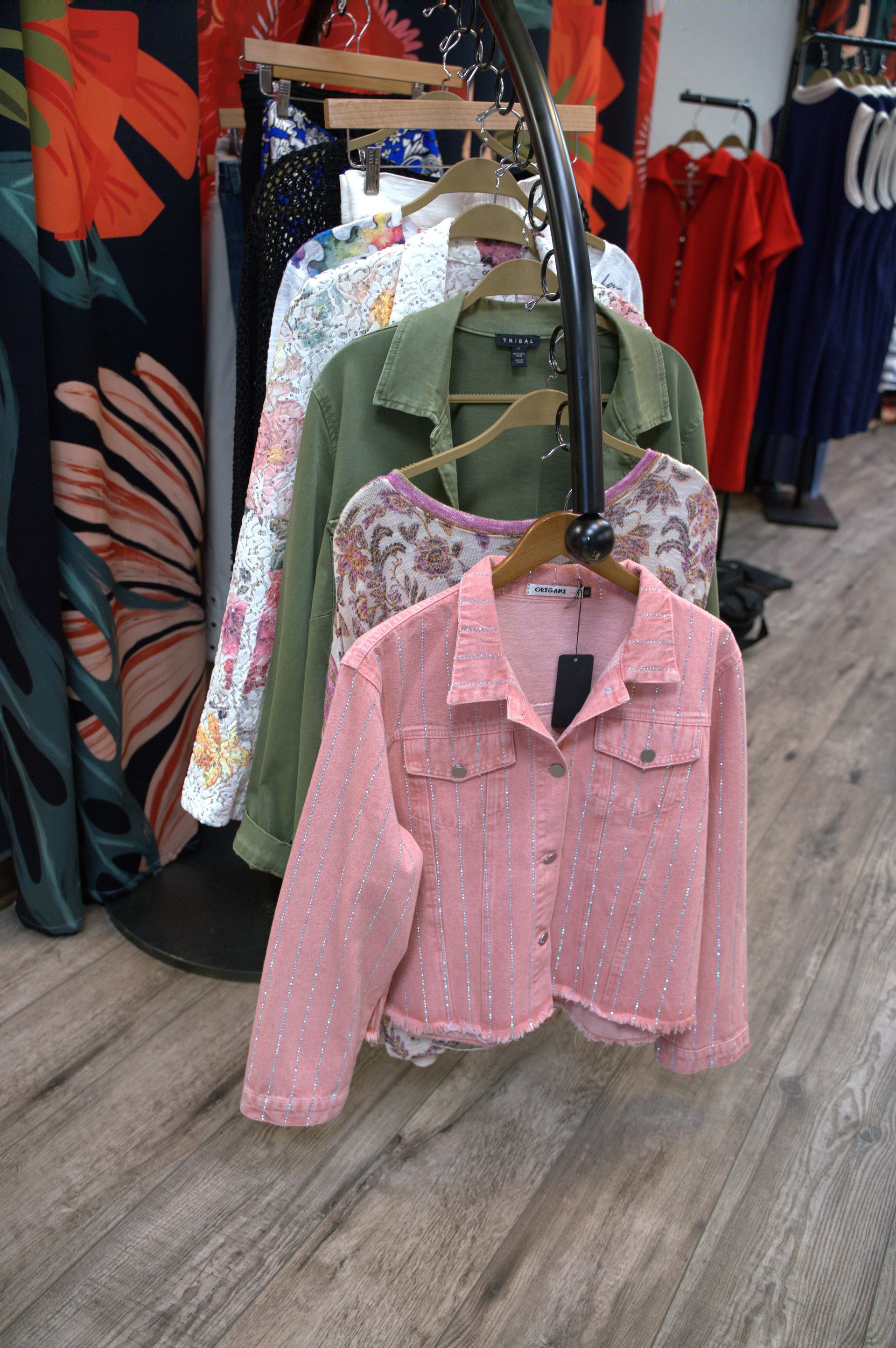 Clothes rack in a boutique, displaying pink, green, and floral jackets. Wooden floor and colorful fabrics in the background.