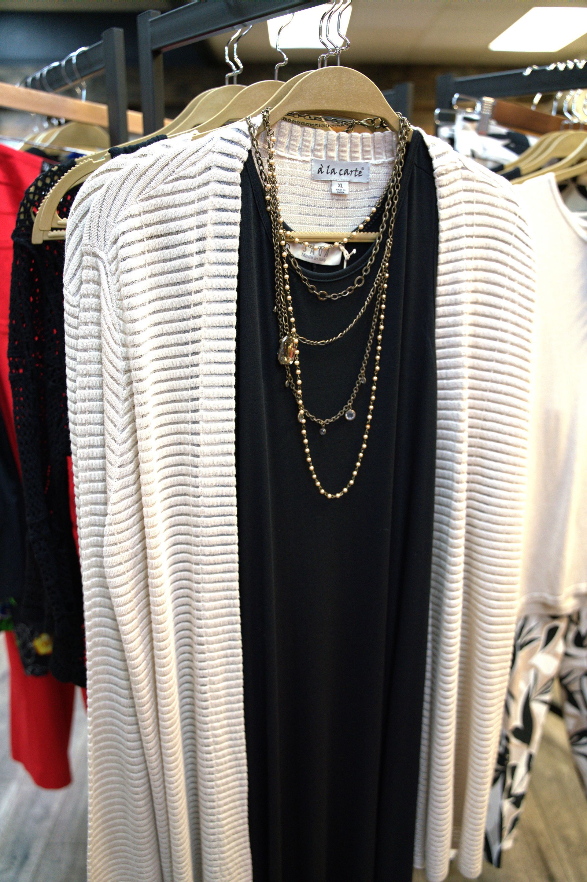 A beige striped cardigan, black dress, and layered gold necklace hang on a clothing rack in a store.
